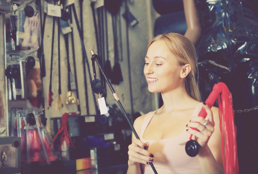 Młoda kobieta jest w sexshopie. Uśmiecha się, w rekach ma bicze