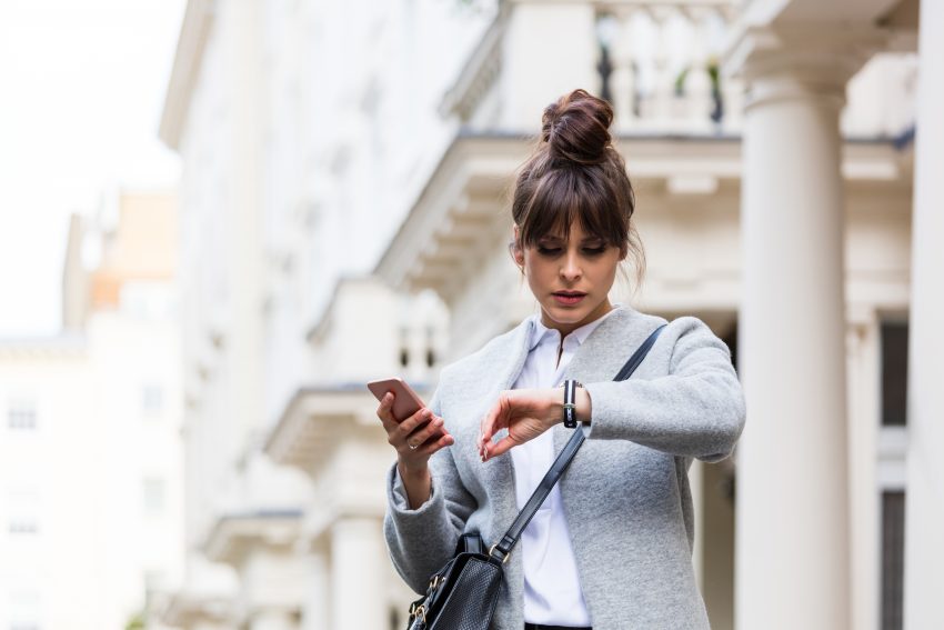 Kobieta uczesana w koka, w jasnym płaszczu i koszulce patrzy na zegarek, trzymając telefon. Widać, że brakuje jej czasu i jest spóźniona