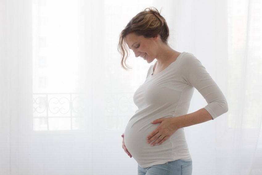 młoda kobieta w ciążyz zamkniętymi oczami trzyma się za brzuch z lekkim uśmiechem. Zdjęcie od boku