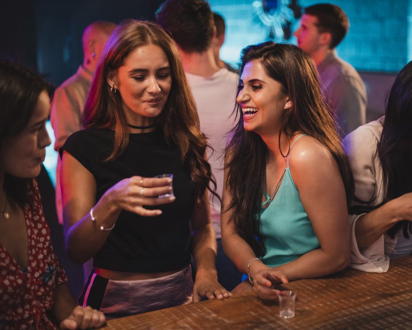 Trzy kobiety stoją przy barze w klubie, jeda pije drinka. Śmieją sie i rozmawiają