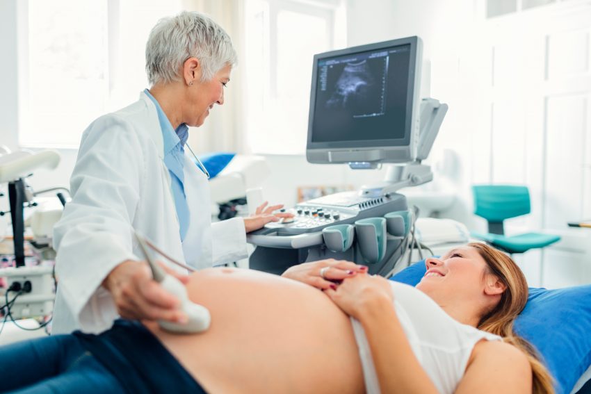 Kobieta w ciąży jest na badaniu u lekarza. Młoda, cieżarna kobieta leży na łóżku w gabinecie i ma odsłonięty brzuch, na którym lekarka trzyma sondę do robienia USG brzucha. Obie panie patrzą na monitor, na którym widać zdjęcie płodu. Lekarka ma fartuch lekarski, siwe włosy, jedną ręką trzyma sondę na brzuchu pacjentki, drugą trzyma na urządzeniu do badania.