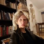 Zuzanna Bartoszek siedzi, na głowie ma zawiązaną tkaninę słomkową jak zwykłą chustę. Za nią regał z książkami oraz szkielet medyczny człowieka