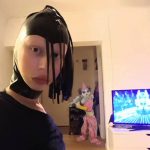 Zuzanna Bartoszek w pokoju, ubrana perwersyjnie w czwarnys strój i czarną skórzaną chustę od głowy do szyi z wycięciem na twarz, na czubku z czarnymi frędzlami. Za nią włączony telewizor oraz wklejony klaun