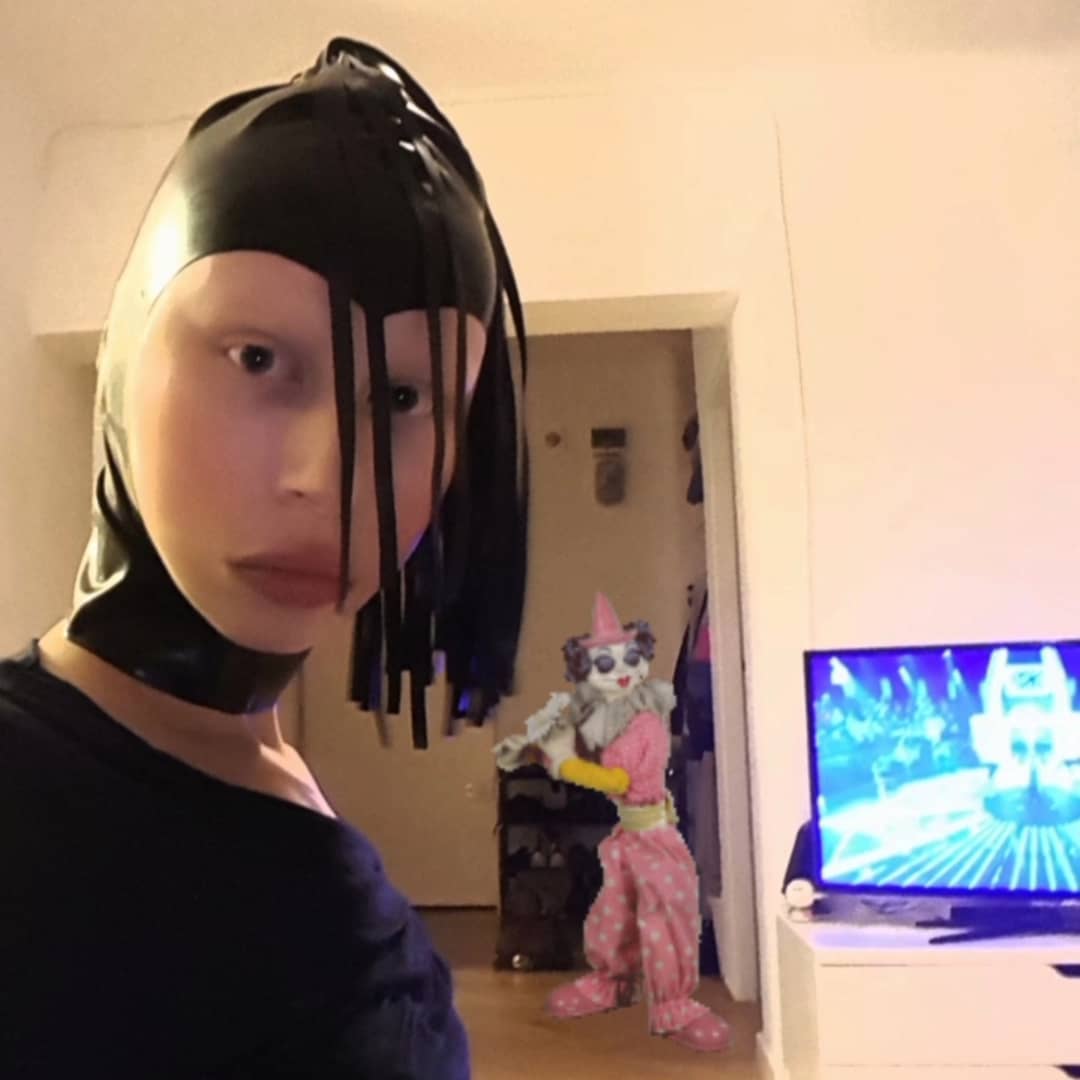 Zuzanna Bartoszek w pokoju, ubrana perwersyjnie w czwarnys strój i czarną skórzaną chustę od głowy do szyi z wycięciem na twarz, na czubku z czarnymi frędzlami. Za nią włączony telewizor oraz wklejony klaun