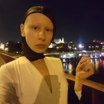 Zuzanna Bartoszek stoi nocą na moście, w tle lampy miasta. Ubrana na biało, na głowie ma czarną kominiarkę z wycięcięm na całą twarz