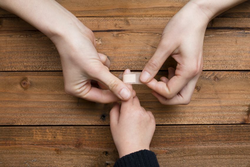 Mama zakleja plaster dziekcu. Na zdjeciu widoczne dłonie na drewnianym stole