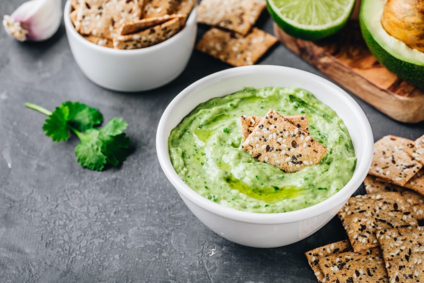 miska z guacamole stoi na czarnym stole, wokół leżą krakersy z sezamem