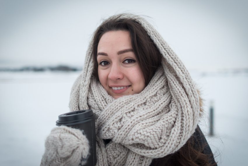 Kobieta stoi na dworze, za nią śnieg. Jest owinięta wełnianym szalem, na ręcę ma grubą rękawiczkę i trzyma kubek z napojem. Kadr do połowy ciała.