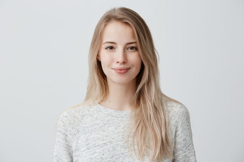 Blondynka w długich włosach w szarej bluzce stoi i uśmiecha się do zdjęcia.