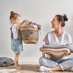 Mama z rócką robią pranie. Mama siedzi ze słożonymi rzeczami a mała dziewczynka przyniosła jej cały kosz ubrań.