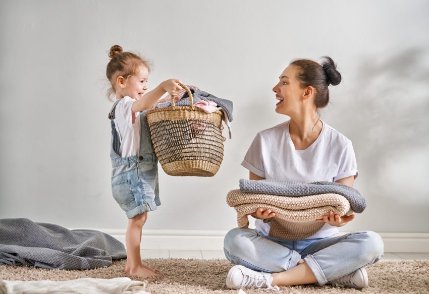 Mama z rócką robią pranie. Mama siedzi ze słożonymi rzeczami a mała dziewczynka przyniosła jej cały kosz ubrań.