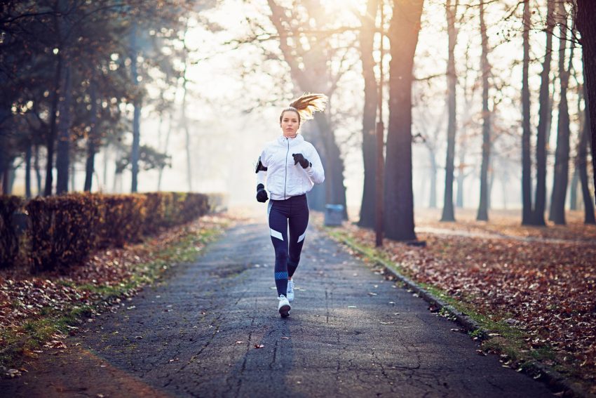Kobieta biegnie ulicą w parku. Jest ubrana w sportowe ciuchy. Dookoła są liście, widać, że jest jesień