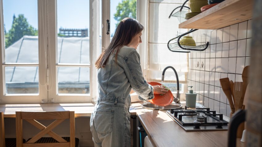 na zdjęciu: kobieta zmywa naczynia w ciasnej kuchni; tekst o tym, jak wpływają na nas nasze mieszkania - Hello Zdrowie