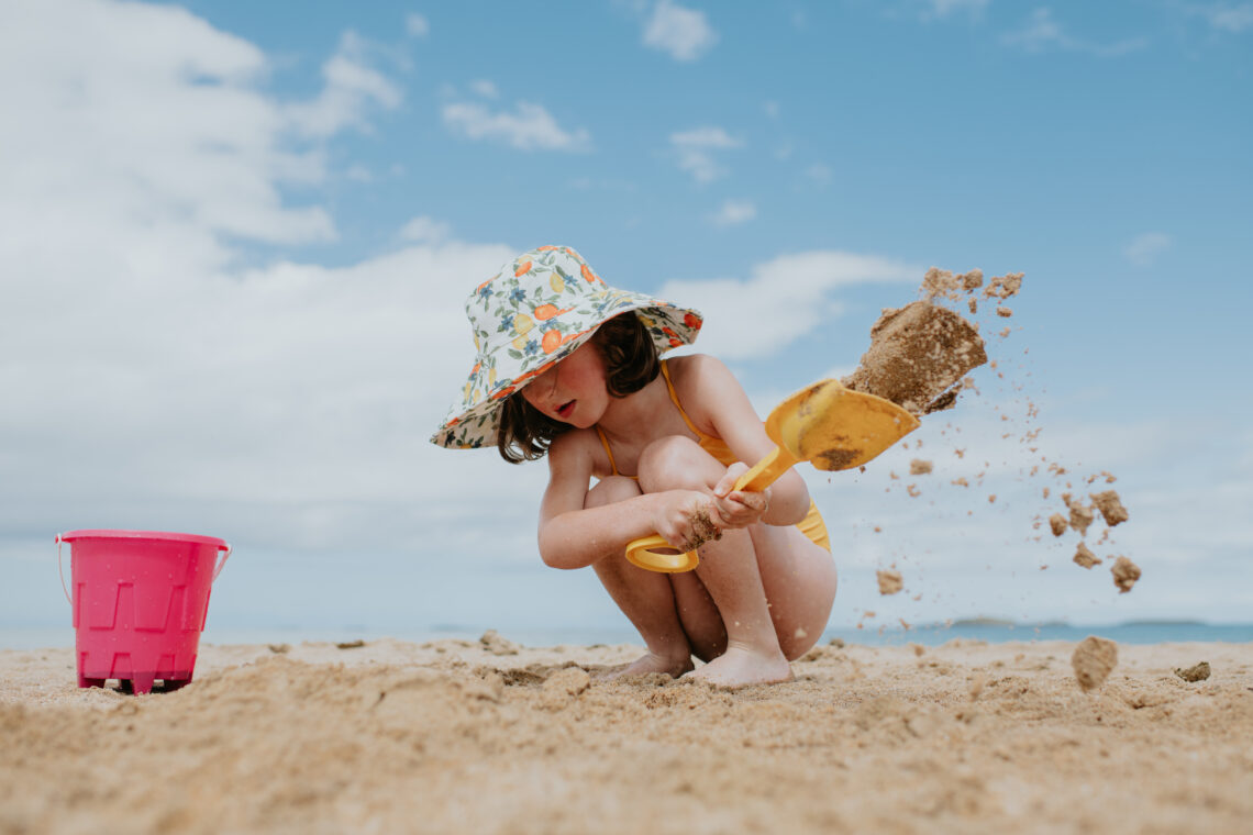 dziewczynka kopie łopatką na plaży, na głowie ma letni kolorowy kapelusz