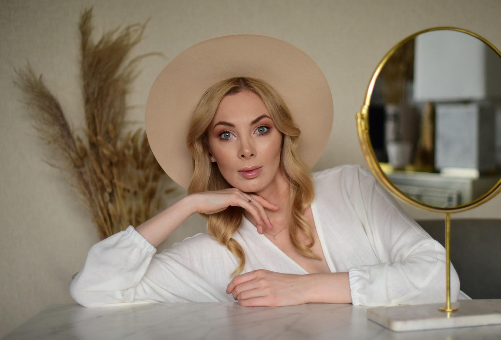Karolina Gliniecka-Hołda, znana jako Charlize Mystery pozuje w beżowym kapeluszu i białej zwiewnej sukience, głowę podpiera ręką