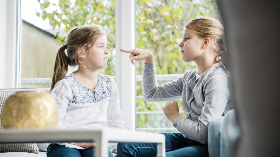 na zdjęciu: dwie dziewczynki siedzą przy oknie, jedna na drugą pokazuje pacem, tekst o przemocy relacyjnej wśród dziewczynek - Hello Zdrowie