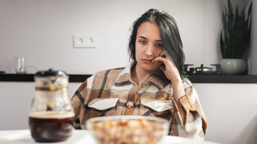 na zdjęciu: zmęczona młoda kobieta siedzi przy kuchennym stole; tekst o produktach, które dodają energii - Hello Zdrowie