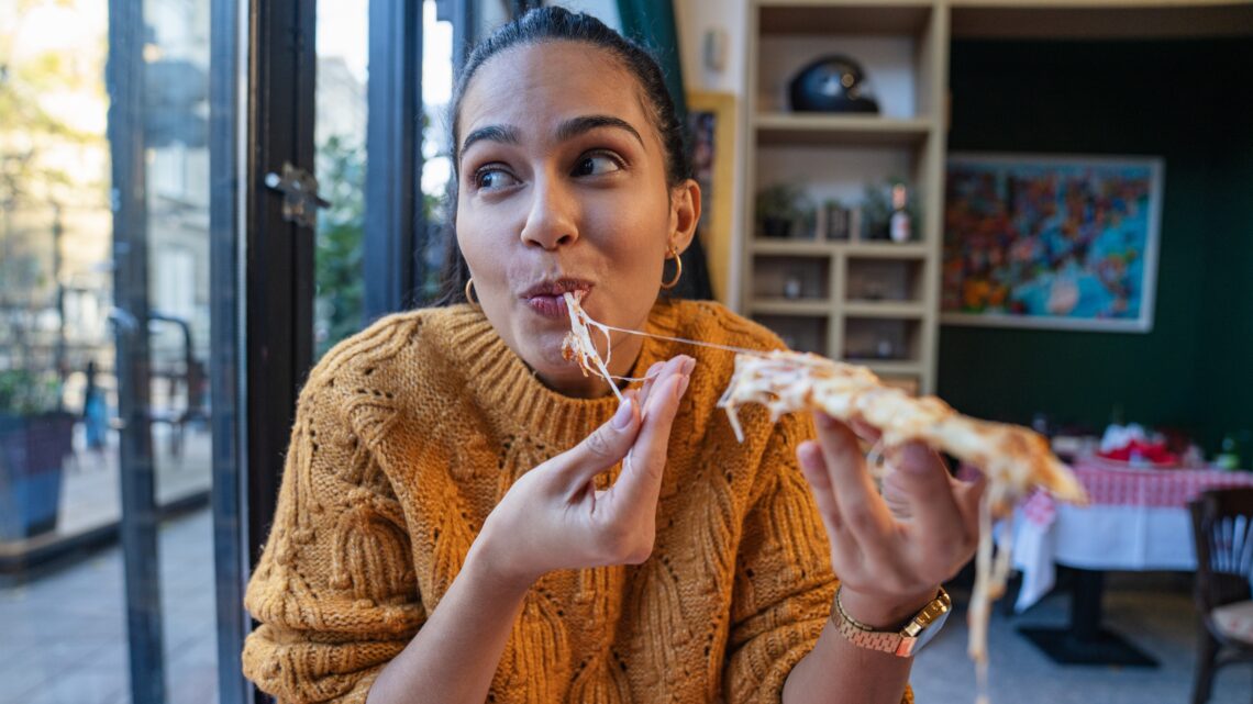 młoda kobieta w żółtym swetrze je pizzę, przytrzymując ciągnący się ser - Hello Zdrowie