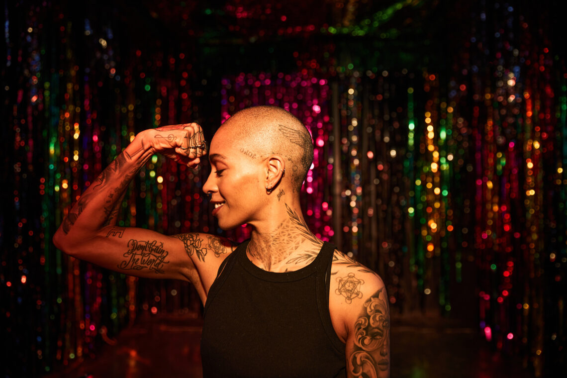 młoda, piękna, łysa, czarnoskóra wytatuowana kobieta pokazuje biceps na tle dyskotekowej kurtyny w klubie - Hello Zdrowie