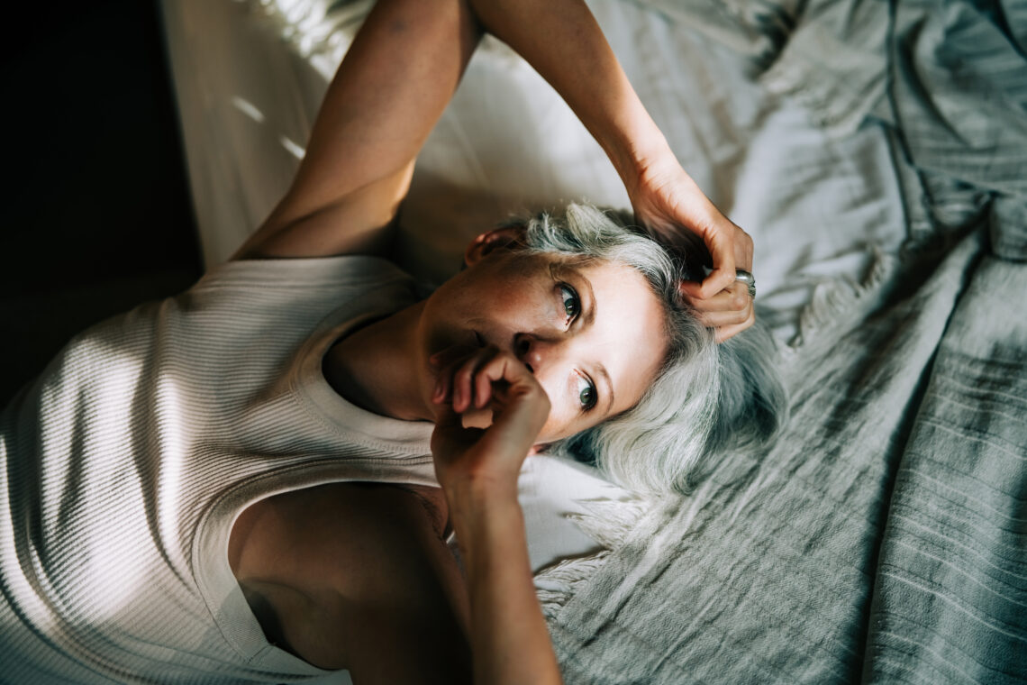 siwowłosa kobieta w białym podkoszulku leży na łóżku w promieniach światła - Hello Zdrowie